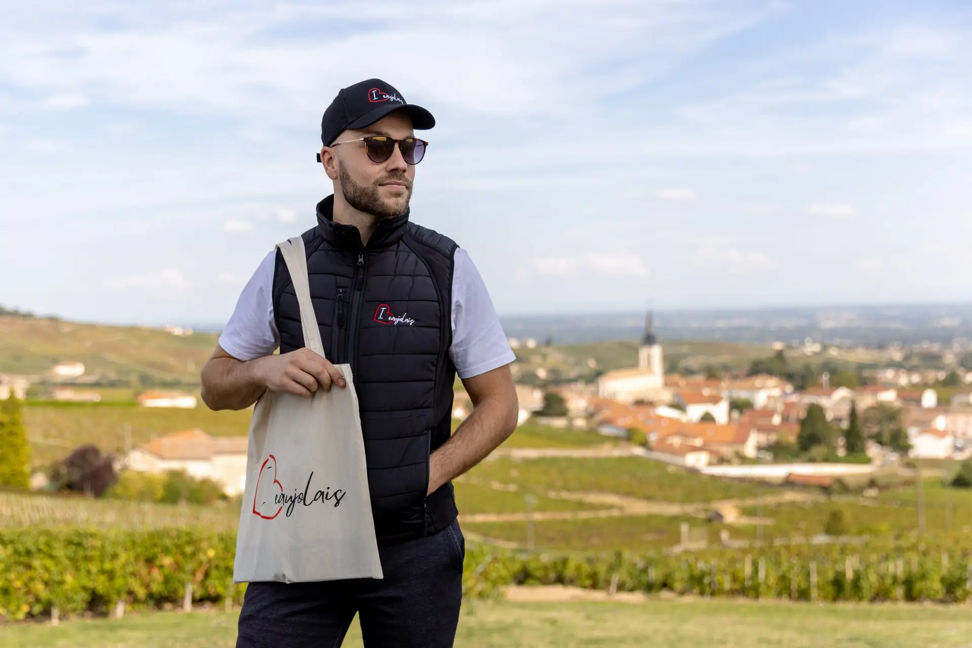 Mannequin homme vêtu d'un tote bag, d'une casquette et d'une doudoune sans manche brodés au nom de la marque I Love Beaujolais. En fond : le paysage de la Madone de Fleurie dans le beaujolais.